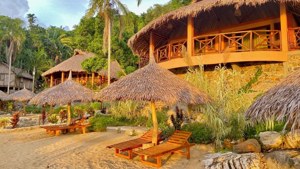 Un resort con estructuras de techo de paja, sillones de madera y una zona de playa arenosa.