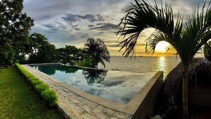 Una piscina infinita con vista al atardecer sobre el océano, rodeada de exuberante vegetación y palmeras.