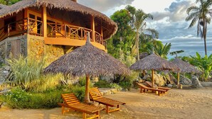 Un resort frente a la playa con construcciones de techo de paja, sillones de madera y exuberante vegetación tropical.