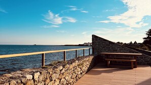 A stone-paved walkway with a wooden bench and a stone wall, overlooking a body of water under a clear blue sky.
