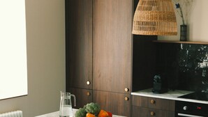 A kitchen with a marble countertop, dark wooden cabinets, a wicker pendant light, and a bowl of fruit. - Maisons Bodenn - Le Penty (Plobannalec-Lesconil)