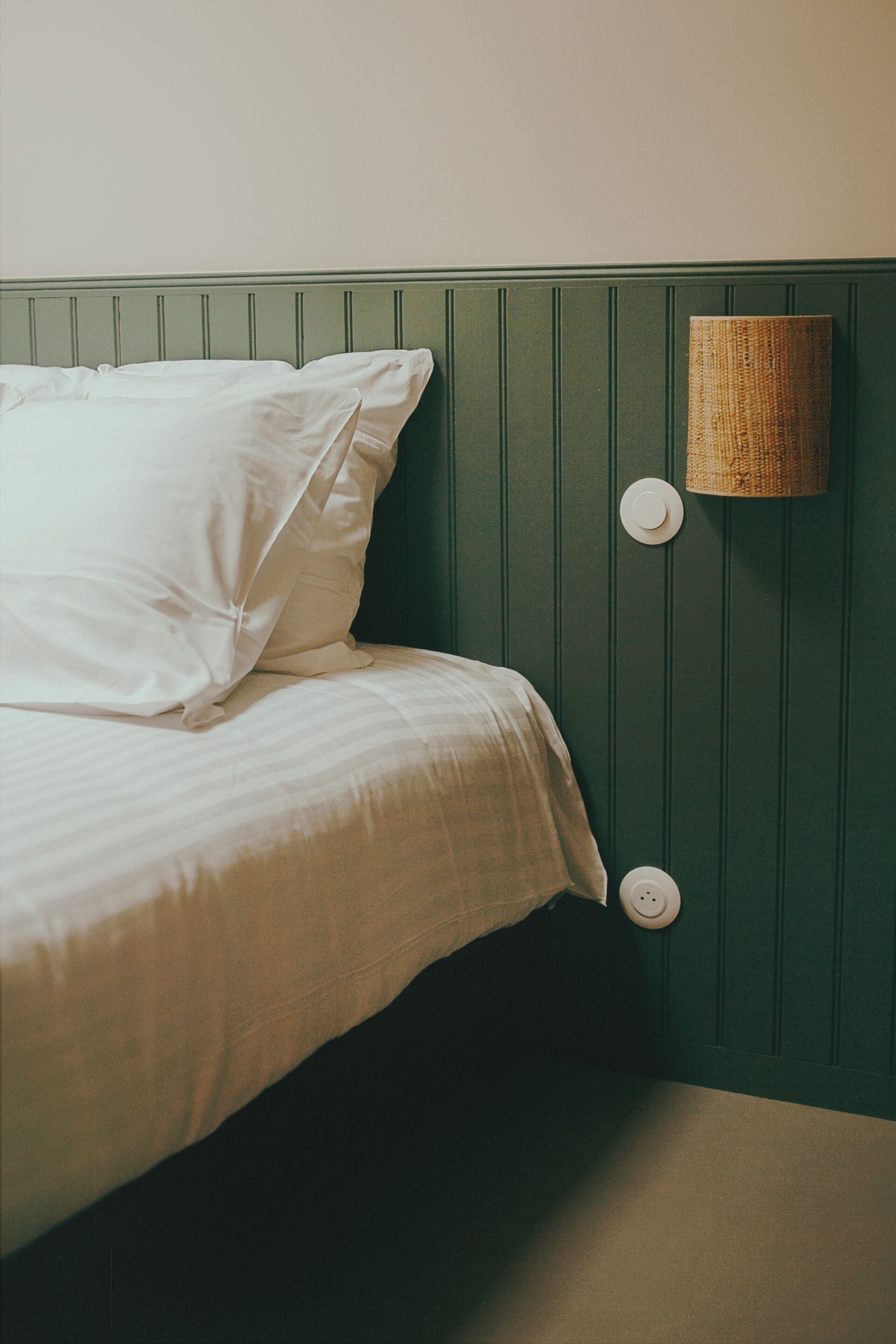 A bed with white bedding in a room with green paneled walls and a woven wall lamp.