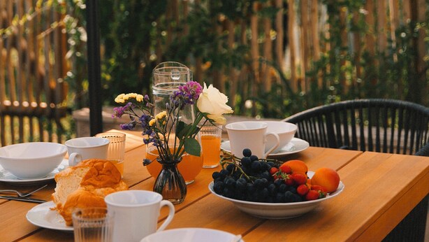 A wooden table set with white cups, saucers, a glass of water, a bowl of fruit, and a small vase with flowers.