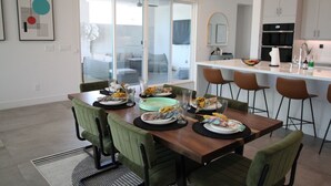 A modern dining area with a wooden table set for six, green upholstered chairs, and a kitchen in the background.