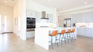 A modern kitchen with white cabinetry, a central island with bar stools, built-in ovens, and a stainless steel refrigerator.