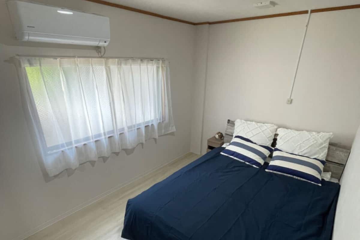 Ein Schlafzimmer mit einem Bett, einem Fenster mit durchsichtigen Vorhängen, einer Klimaanlage und einem Holzboden.