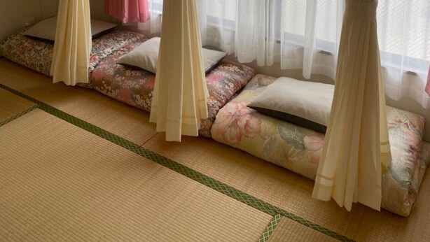 Une pièce au sol recouvert de tatamis, avec un futon et une literie à motifs floraux, et des rideaux légers.