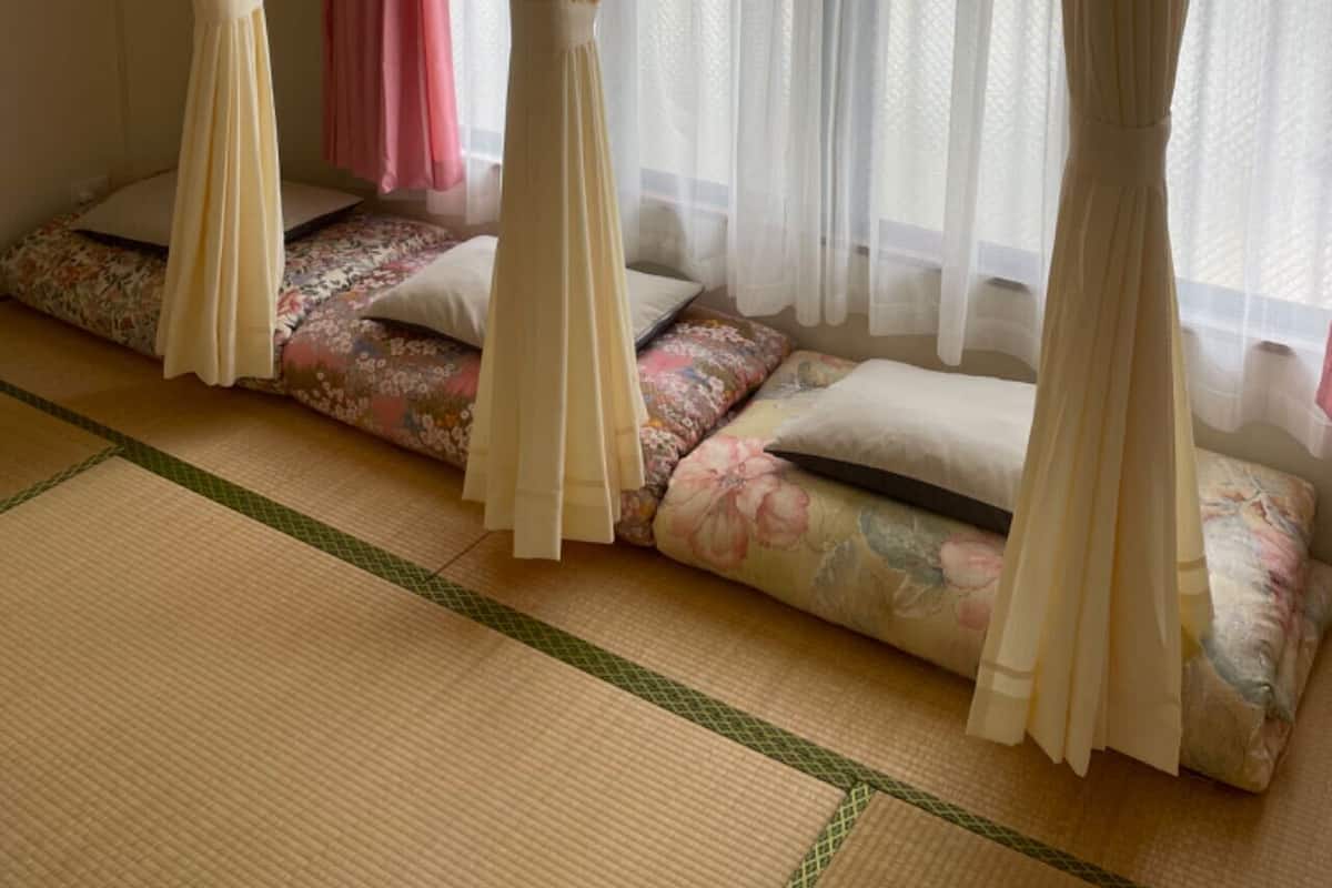 Ein Zimmer mit Tatami-Boden, einem Futonbett mit floralem Bettzeug und leichten Vorhängen.