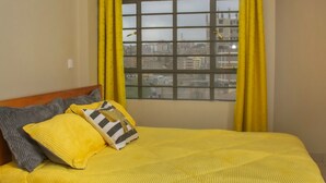 A bed with a yellow bedspread and pillows, a window with yellow curtains, and a view of buildings outside. - Lux Suites Clay City Heights Kasarani (Nairobi)