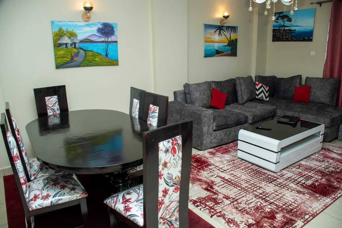 A living room with a grey sofa, red cushions, a round dining table with floral chairs, and three paintings on the walls.
