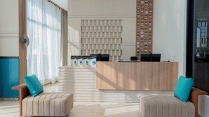 A modern reception area with a wooden reception desk, two beige sofas with teal cushions, a large window with sheer curtains, and a wall-mounted shelving unit. - Sunway Tabuk Hotel (Tabuk)