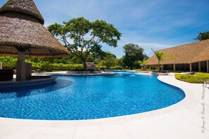 A swimming pool with a curved edge, surrounded by thatched-roof structures and lush greenery. - Ciudad Del Mar 126 3 Bedroom Condo by RedAwning (Jacó)