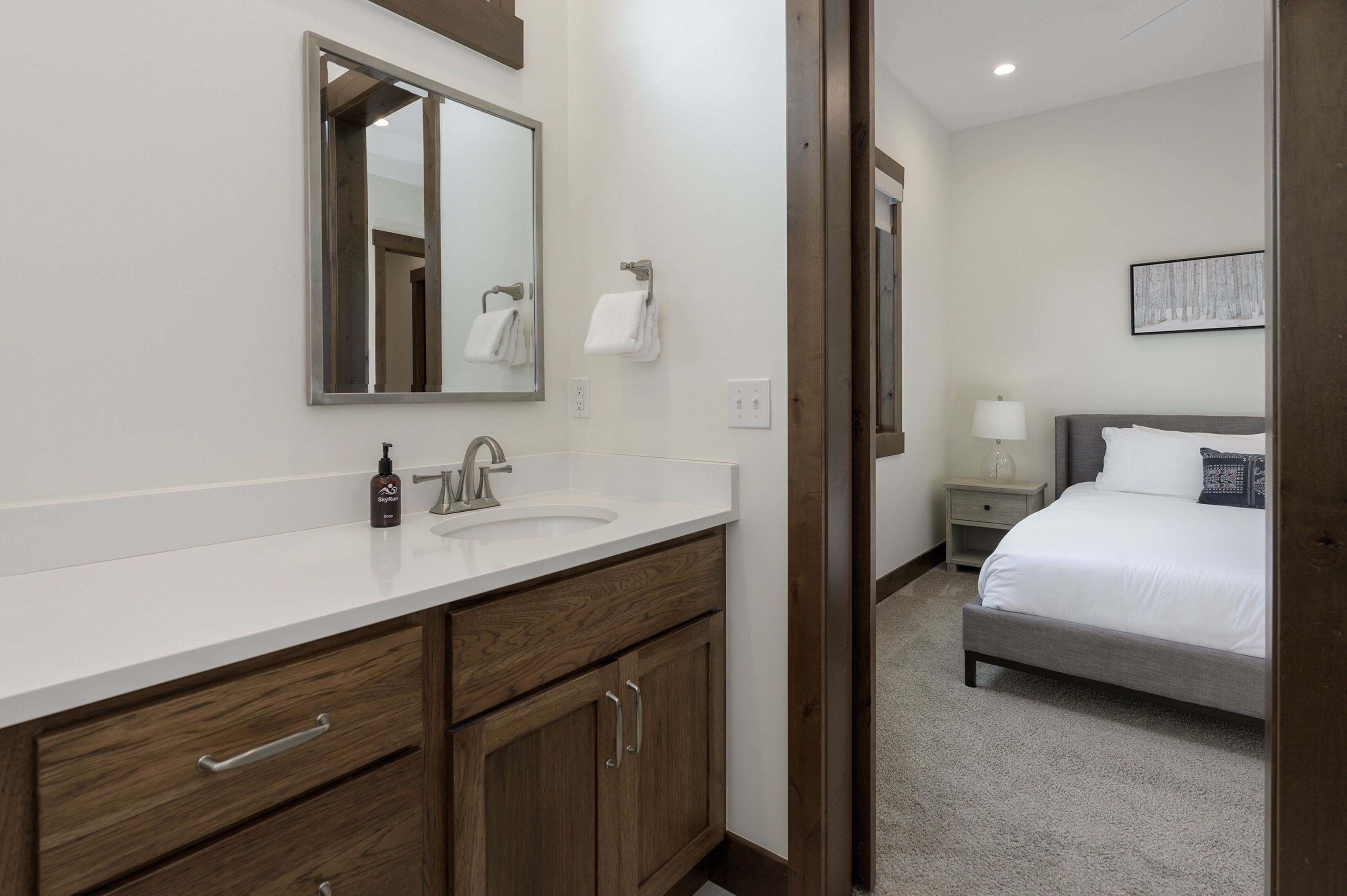 A modern hotel bathroom with a large mirror, wooden vanity, and a separate bedroom with a bed and nightstand.