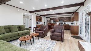 A spacious living area with a green sofa, two armchairs, and a coffee table. A kitchen with wooden cabinets and modern appliances is visible in the background. - Becca's Place 3 Bedroom Home by RedAwning (Pigeon Forge)