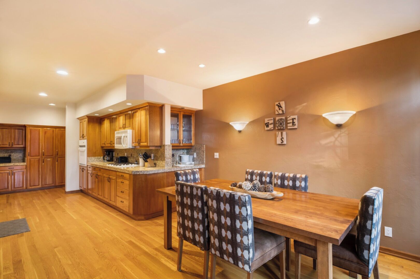 A modern kitchen with wooden cabinets and a central island, a dining area with a wooden table and patterned chairs, and a wall with decorative lights and framed artwork.