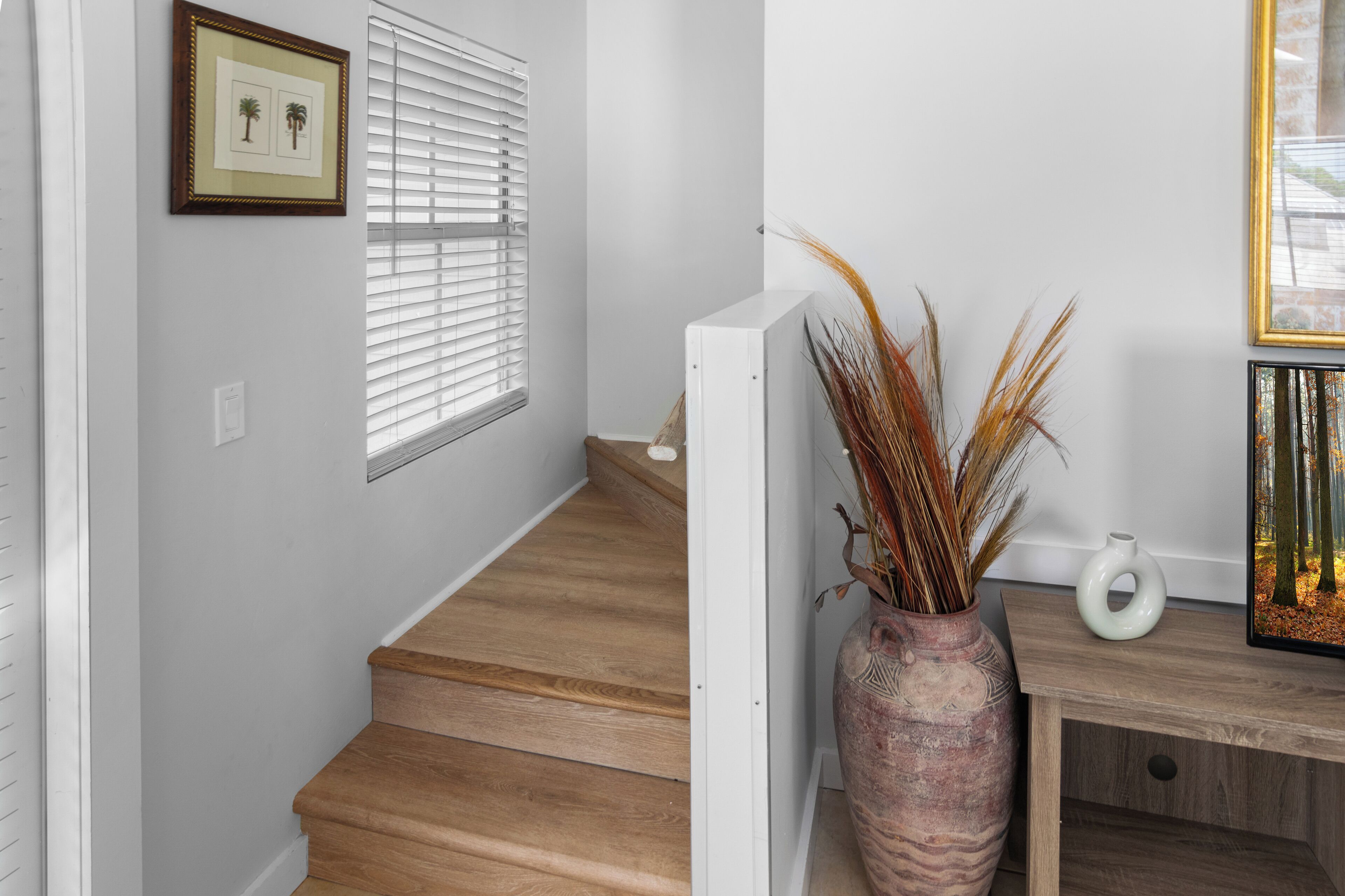Eine Treppe mit Holzdielen, eine weiße Wand, ein gerahmtes Bild, ein Fenster mit Jalousien, eine Vase mit Trockenpflanzen und ein kleiner Tisch mit einer Vase.