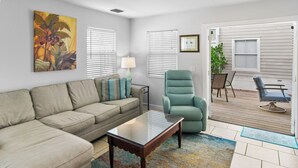 A living room with a beige sectional sofa, a glass coffee table, a teal armchair, and a wooden deck with outdoor furniture. - THALIA'S RETREAT @ Duval Square 2bd/2ba townhome (Key West)