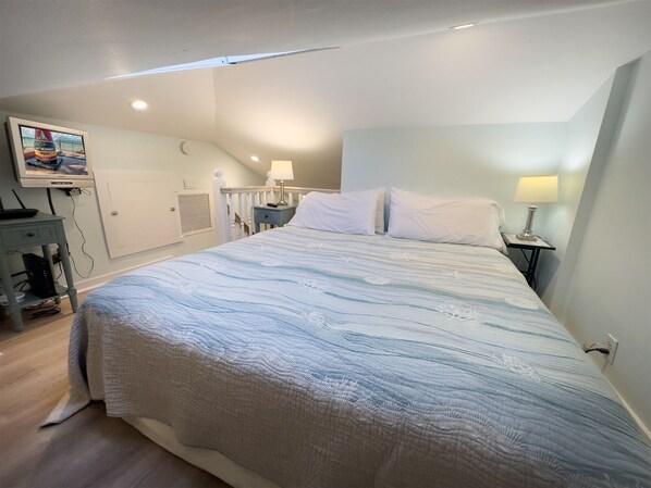 A bedroom with a bed, two bedside tables with lamps, a desk with a computer, and a TV on the wall.