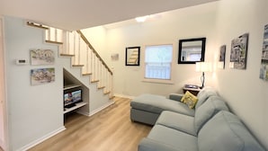 A modern living room with a grey sofa, a TV on a stand, a staircase, and framed pictures on the walls. - Rent at PORCHSIDE PARADISE - Steps to Duval (Key West)