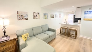 A living room with a light green sofa, a wooden side table, and a lamp. There are two wooden stools and a kitchen area with a sink and countertop. - Rent at PORCHSIDE PARADISE - Steps to Duval (Key West)