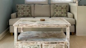 Une pièce chaleureuse avec un ventilateur de plafond, deux fenêtres équipées de stores, un canapé agrémenté de coussins à motifs et une table basse rustique.