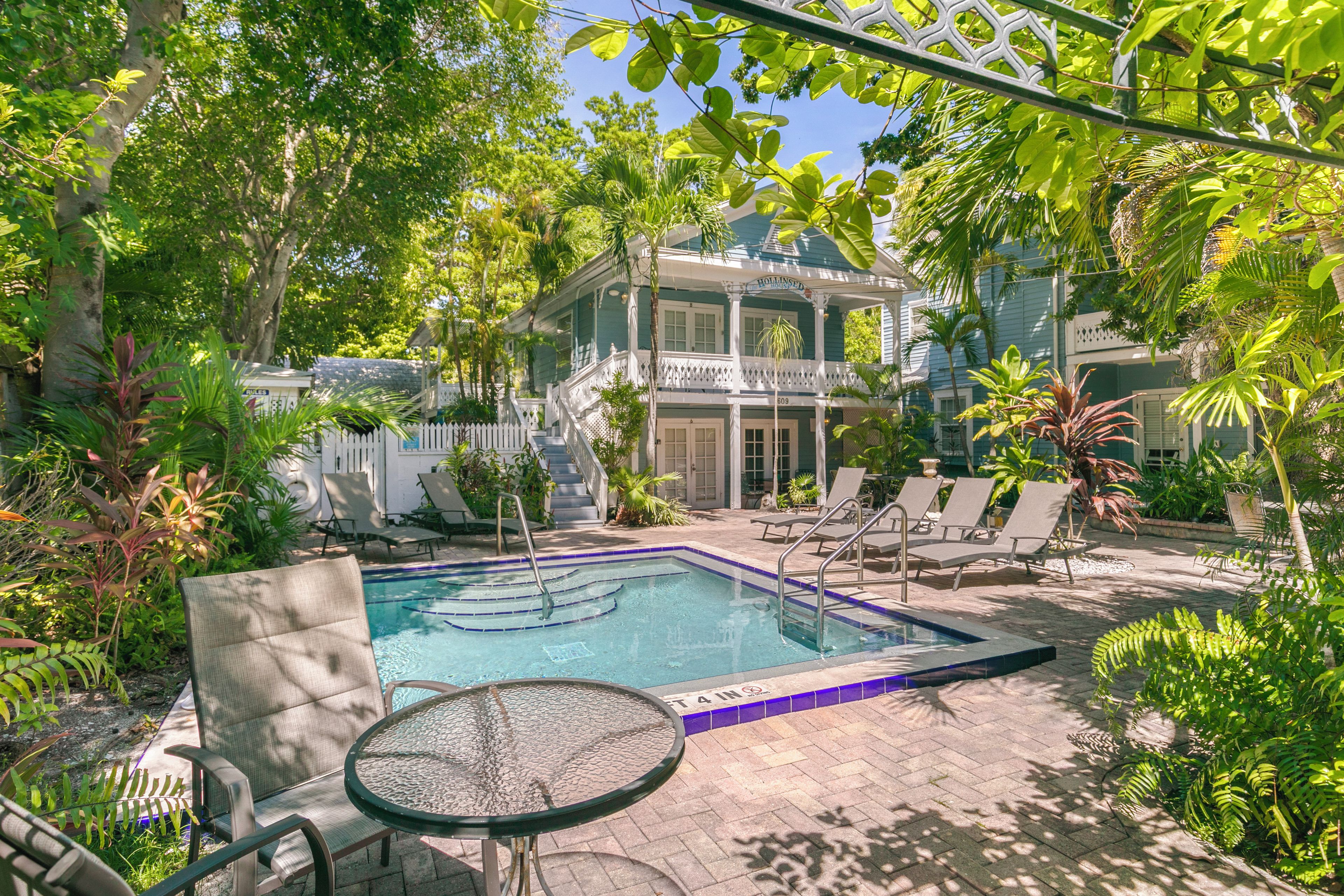 A pool area with lounge chairs and a table, surrounded by lush greenery and a building with a porch.