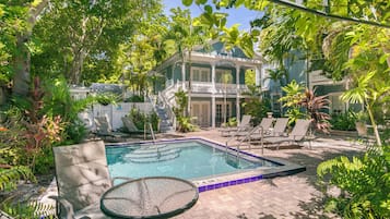 A pool area with lounge chairs and a table, surrounded by lush greenery and a building with a porch.