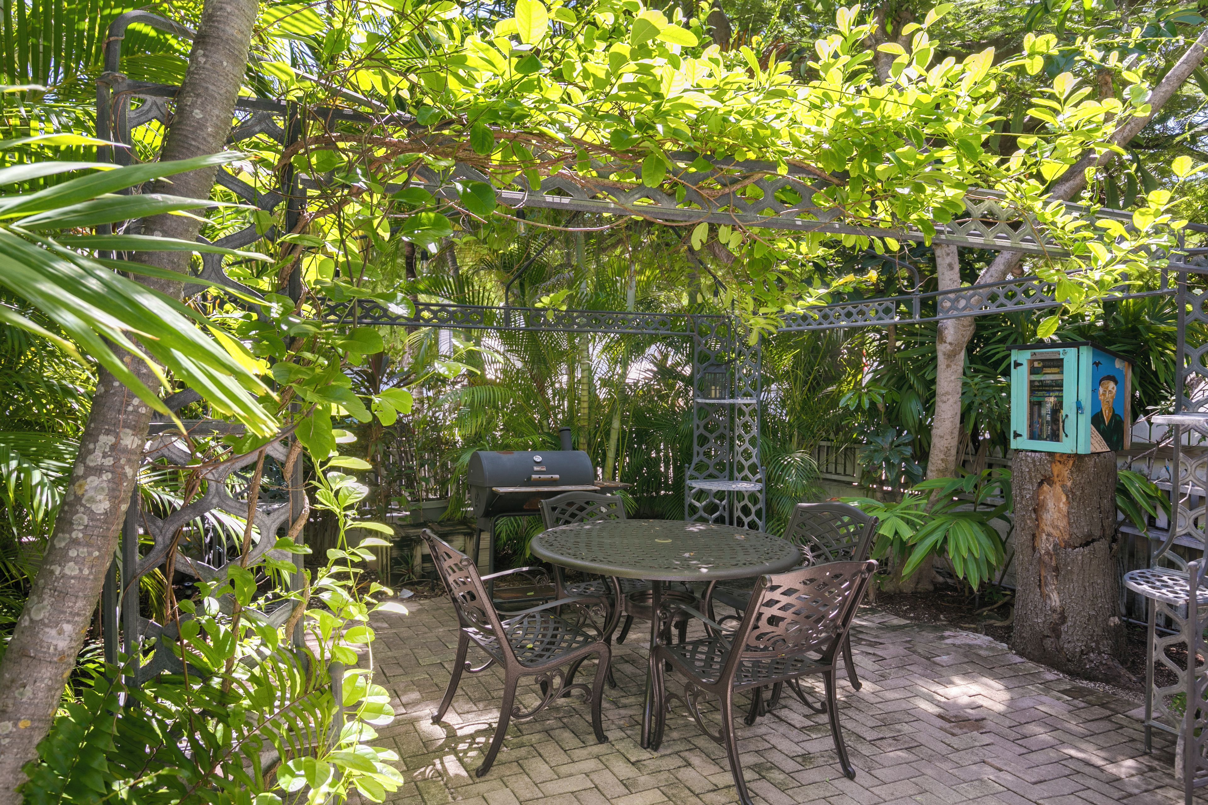 An outdoor patio with a round table and chairs, surrounded by lush greenery and a metal grill.