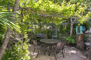 An outdoor patio with a round table and chairs, surrounded by lush greenery and a metal grill. - Hollinsed House Studio - Great Location!! (Key West)