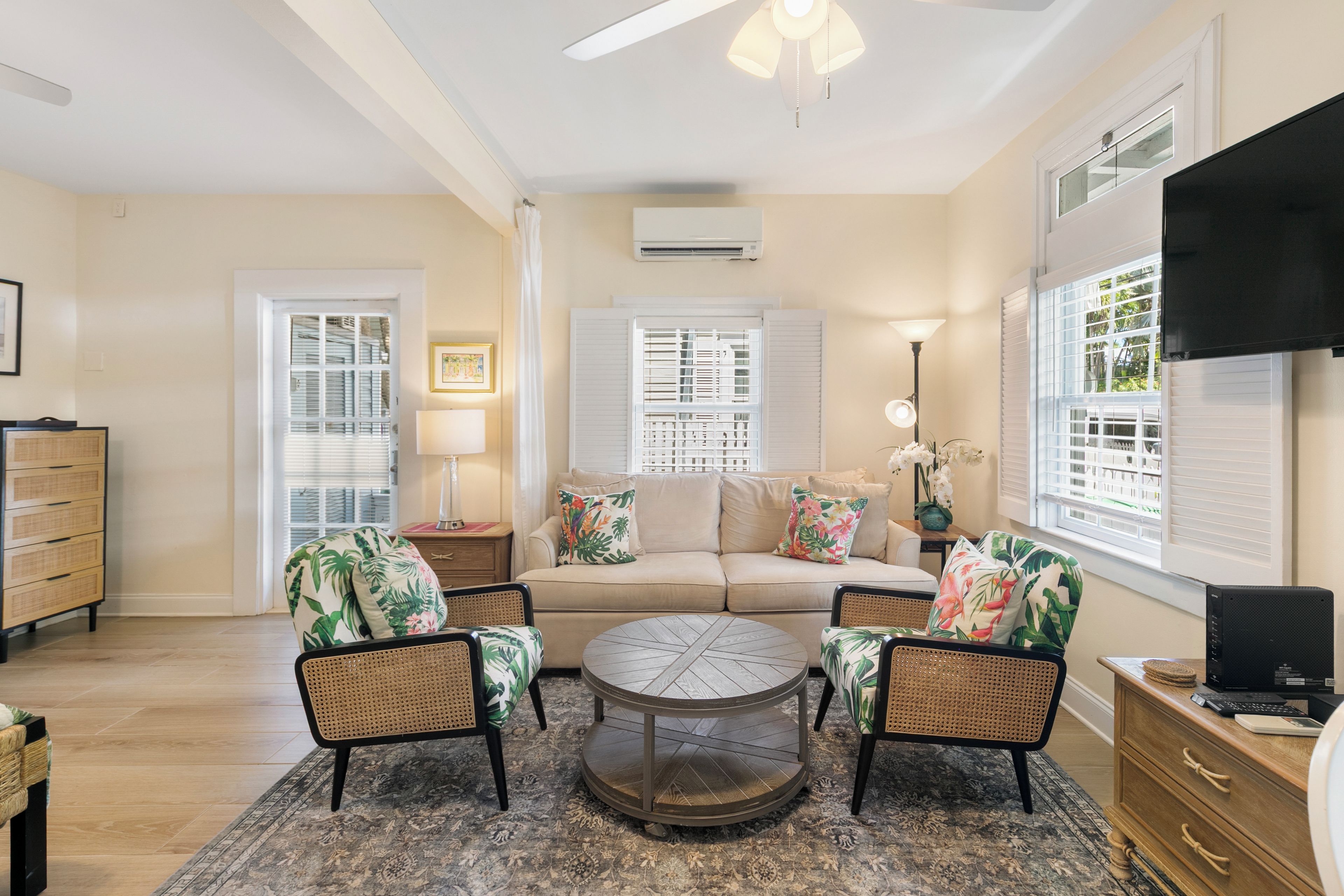 A living room with a beige sofa, two rattan chairs, a round coffee table, and a flat-screen TV mounted on the wall.