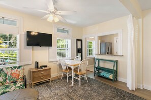 A room with a ceiling fan, a flat-screen TV mounted on the wall, a dining table with chairs, a small wooden cabinet, and a green console table with towels.