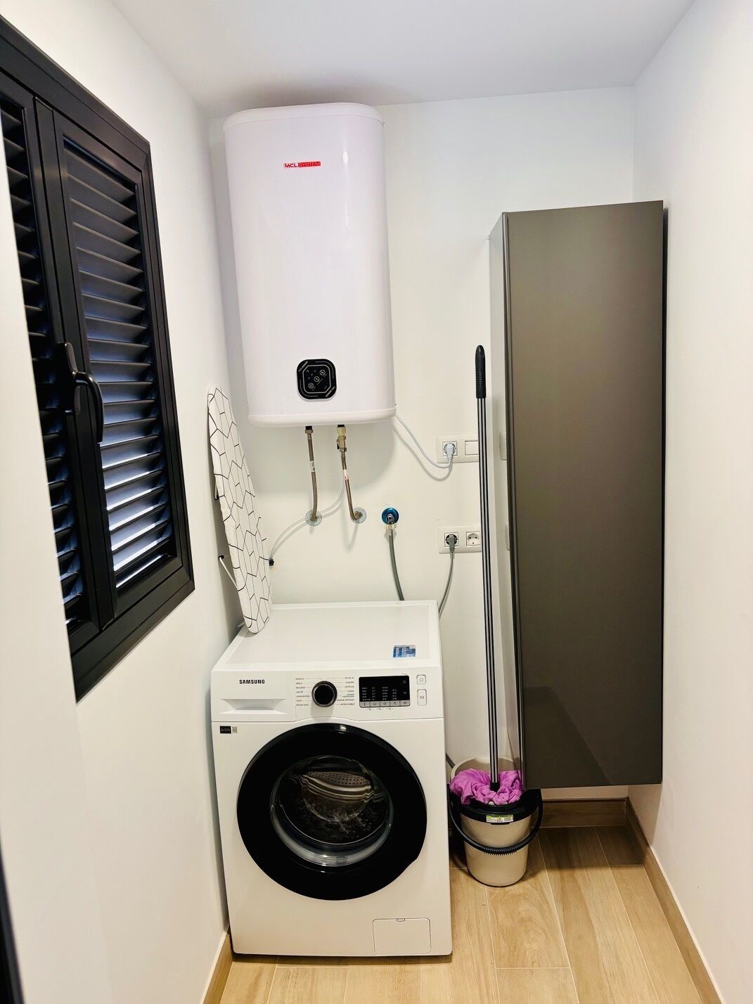 Een kleine wasruimte met een witte wandwaterverwarmer, een Samsung wasmachine en een grijze kast.