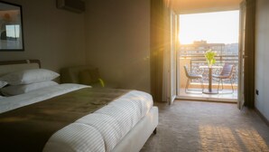 A hotel room with a bed, a chair, and a balcony with a view of buildings. - Downtown Apartments (Pristina)