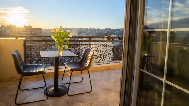 A balcony with a table and chairs, a vase of flowers, and a view of buildings in the distance. - Downtown Apartments (Pristina)