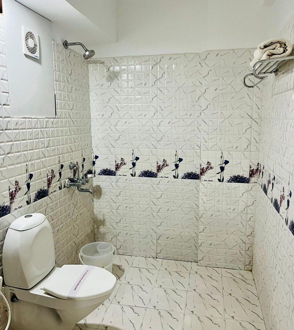Une salle de bain avec des toilettes blanches, une douche équipée d’un pommeau de douche amovible et un porte-serviettes.