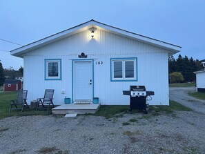 A small, single-story building with a white exterior and blue trim. It features a front door, two windows, and a small porch with two chairs. A barbecue grill is positioned outside the building. - Cozy home with lots of nature trails with ocean views close to all amenities.  (Port au Port East)