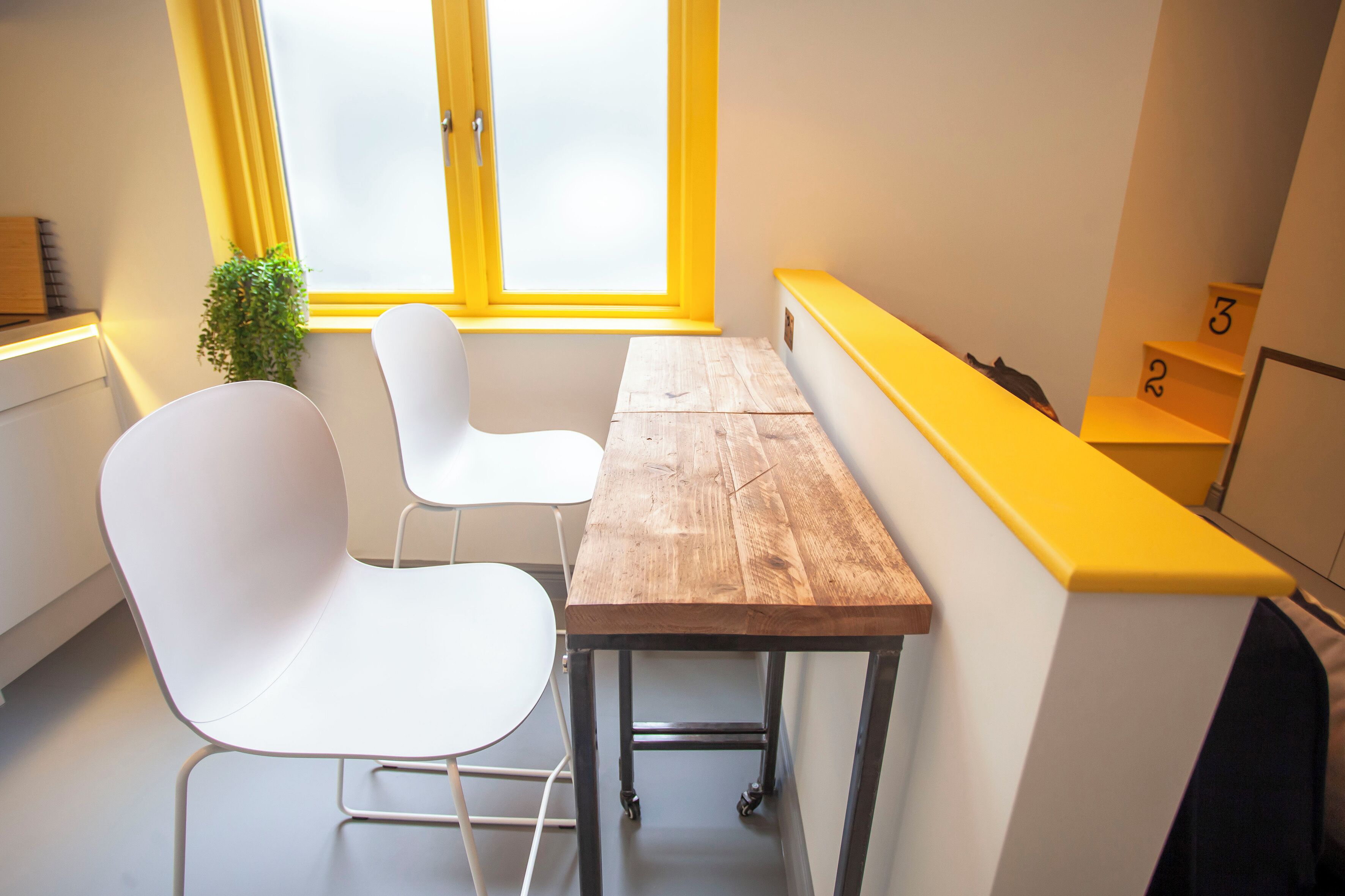 A modern room with a wooden table, white chairs, a yellow bench, and a window with yellow curtains.