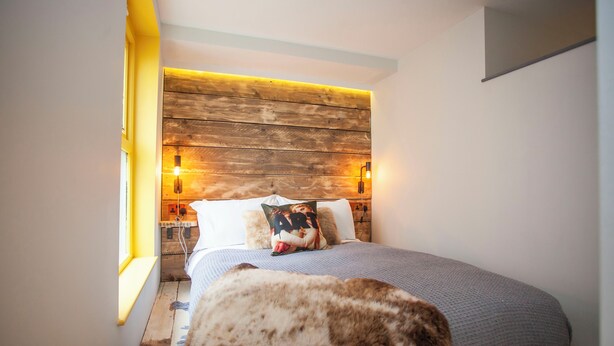Un dormitorio con cabecera de madera, cama con manta gris y almohada con diseño floral, y un gato sobre la cama.