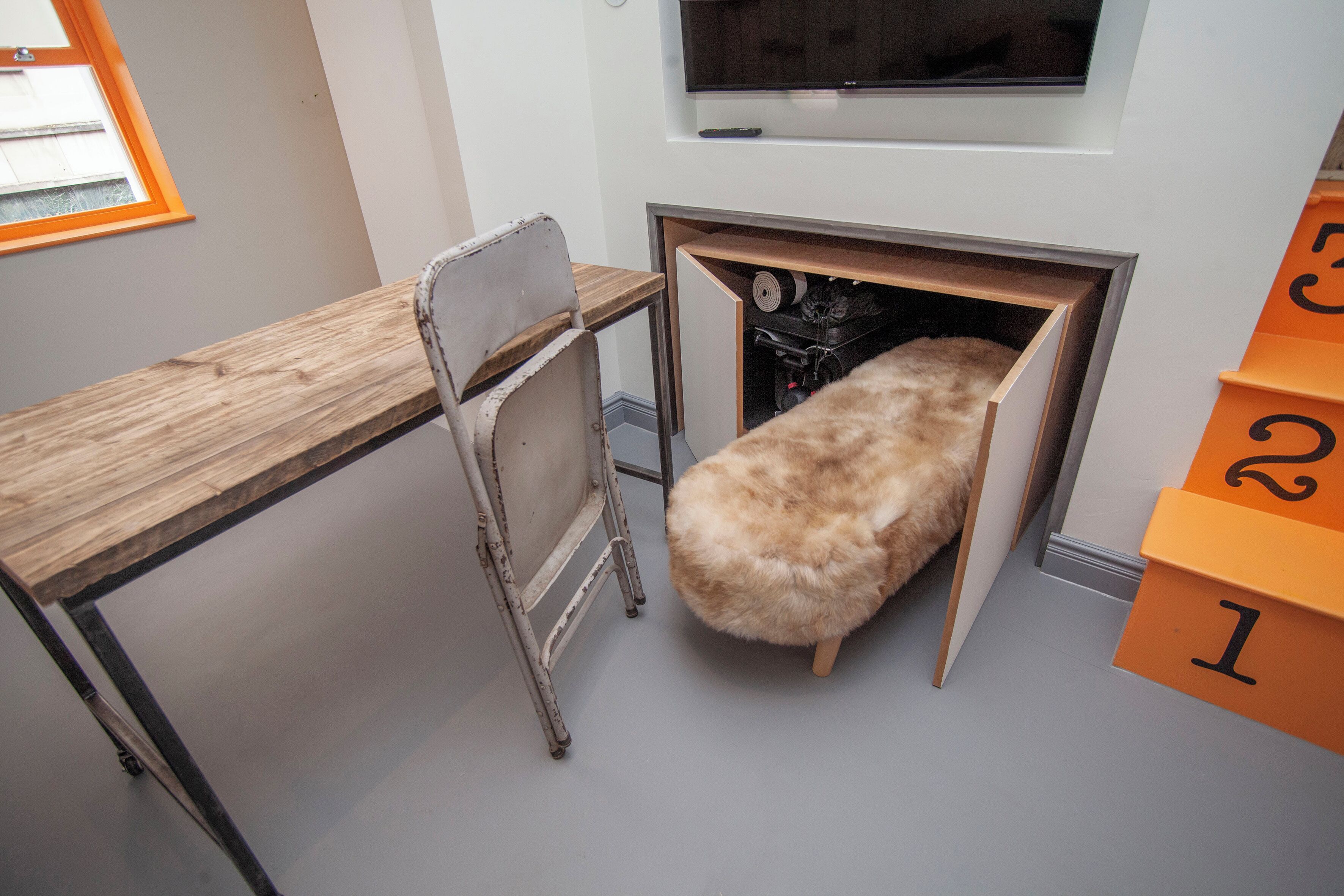 A small room with a wooden desk, a chair, a bench, and a television mounted on the wall.