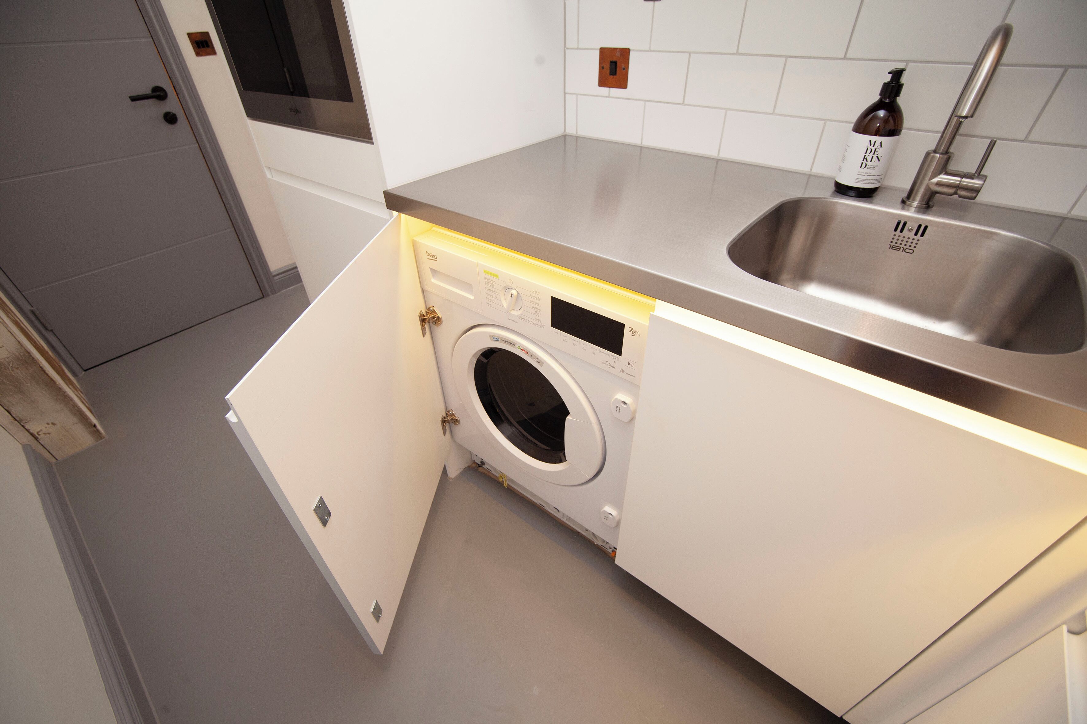 Une cuisine compacte équipée d’un lave-linge intégré, d’un évier en acier inoxydable et d’un flacon de savon liquide pour les mains.