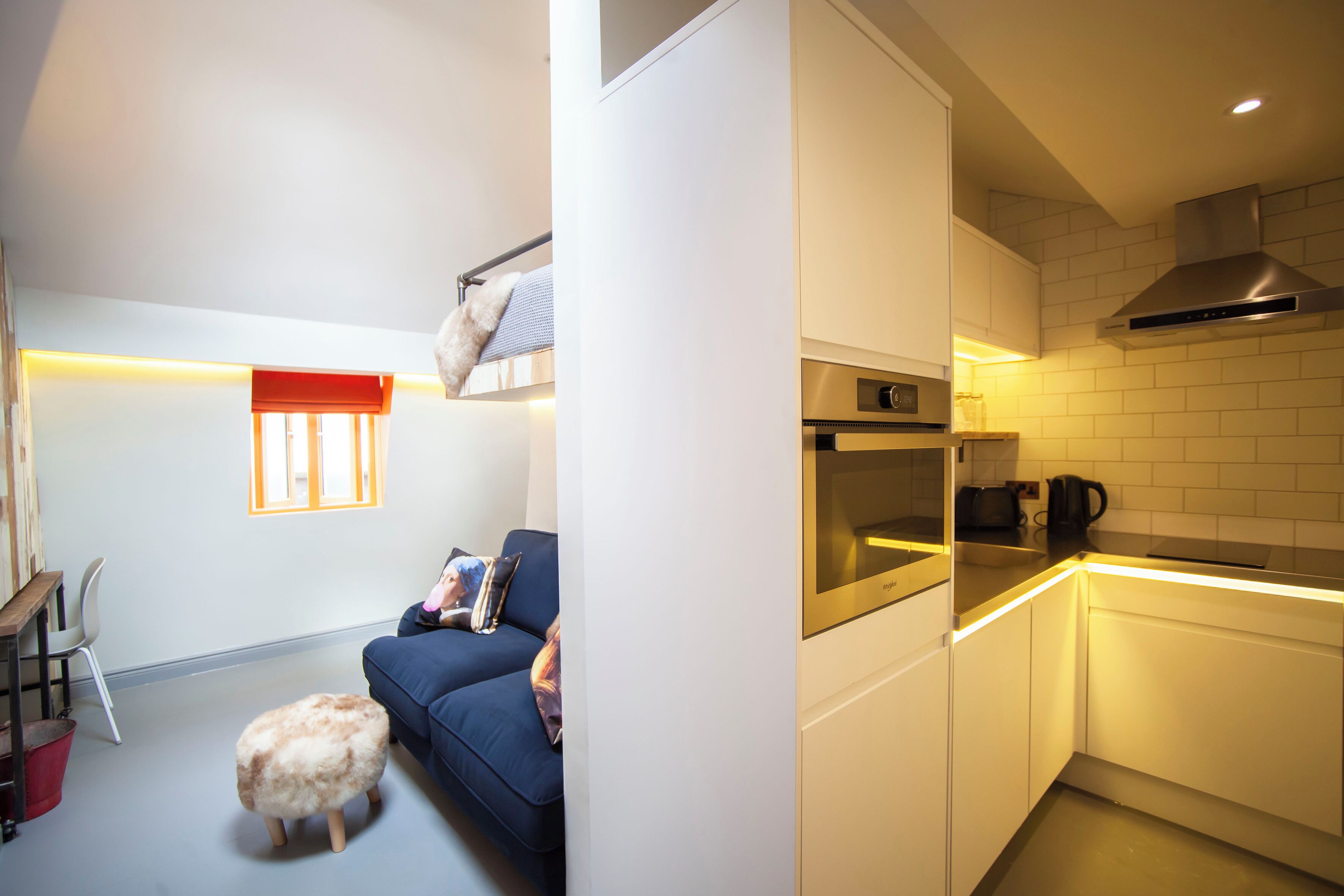 Un espacio habitable compacto con kitchenette, un sofá, un taburete pequeño y una ventana con persianas.