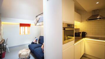 Un espacio habitable compacto con kitchenette, un sofá, un taburete pequeño y una ventana con persianas.