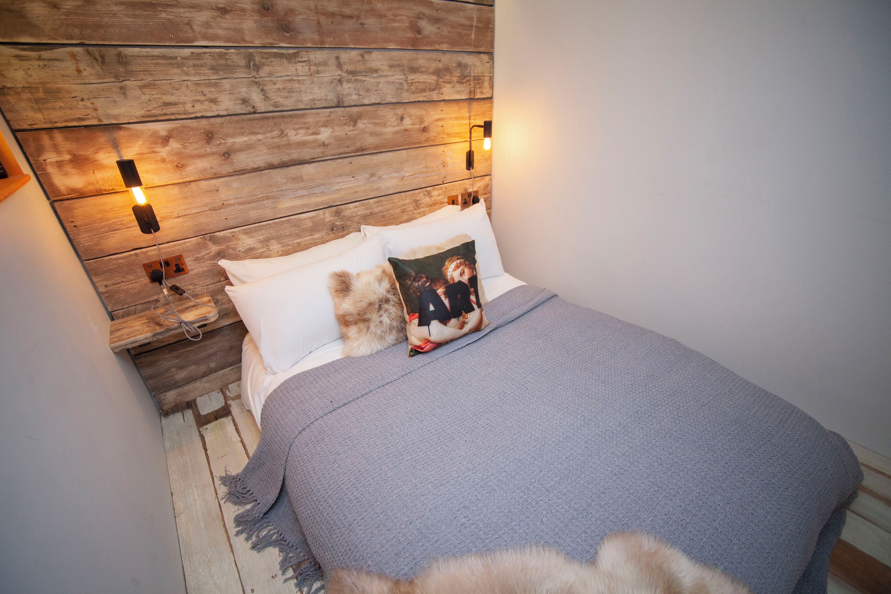 Camera da letto con testiera in legno, coperta grigia e cuscino con un'immagine.