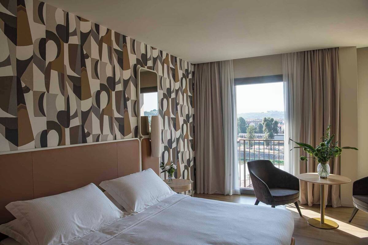 A hotel room with a large bed, a modern abstract wall design, a view of the city through the window, and a small round table with a vase of flowers.