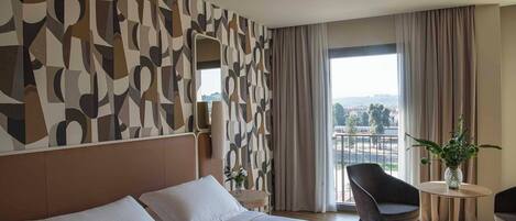 A hotel room with a large bed, a modern abstract wall design, a view of the city through the window, and a small round table with a vase of flowers.