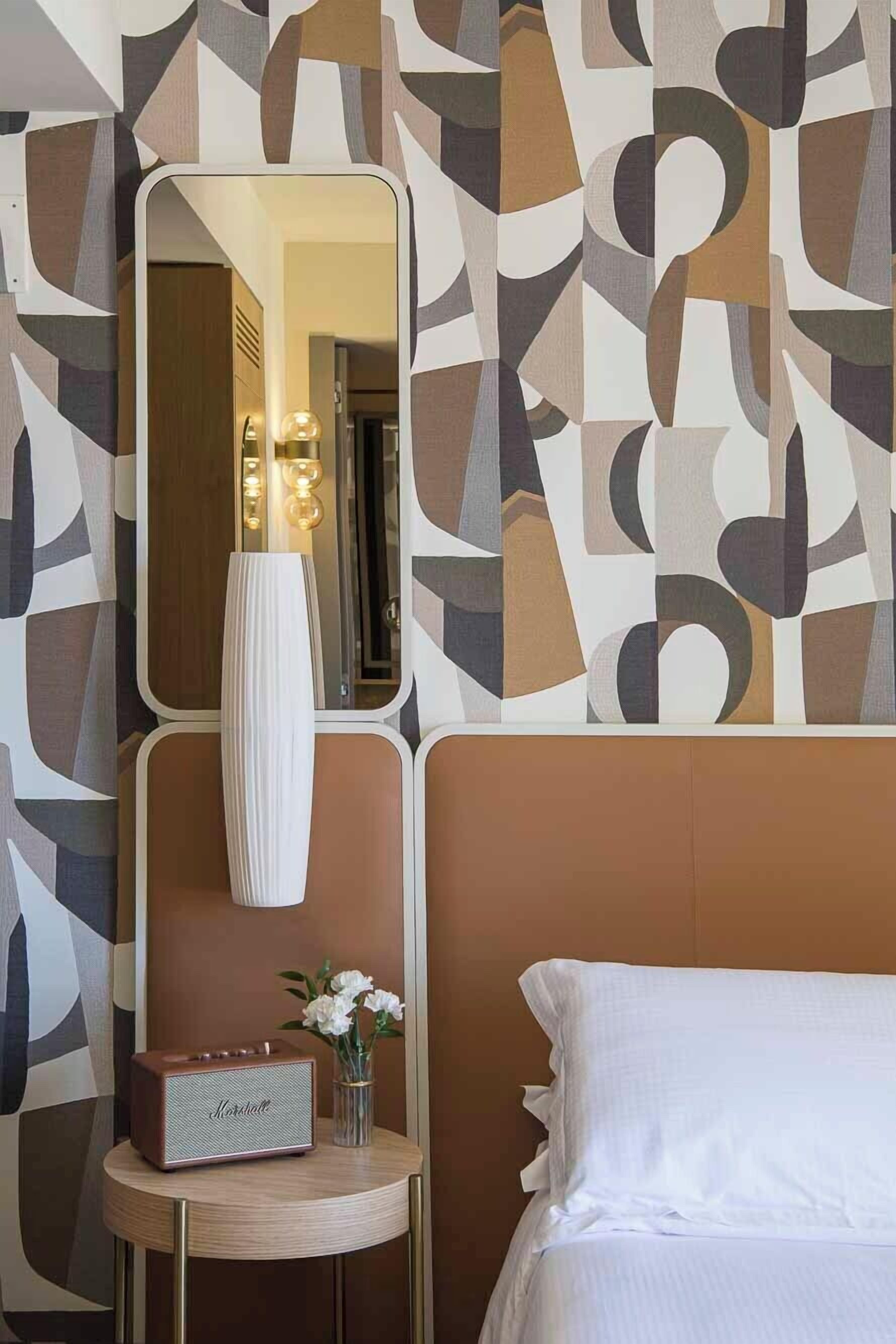 Habitación de hotel con diseño moderno, que incluye un espejo grande, una mesita de noche con lámpara, una radio y un florero con flores.