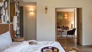 A hotel room with a bed, bedside table, lamp, and a view into the dining area. - Columbus Urban Resort (Florence)