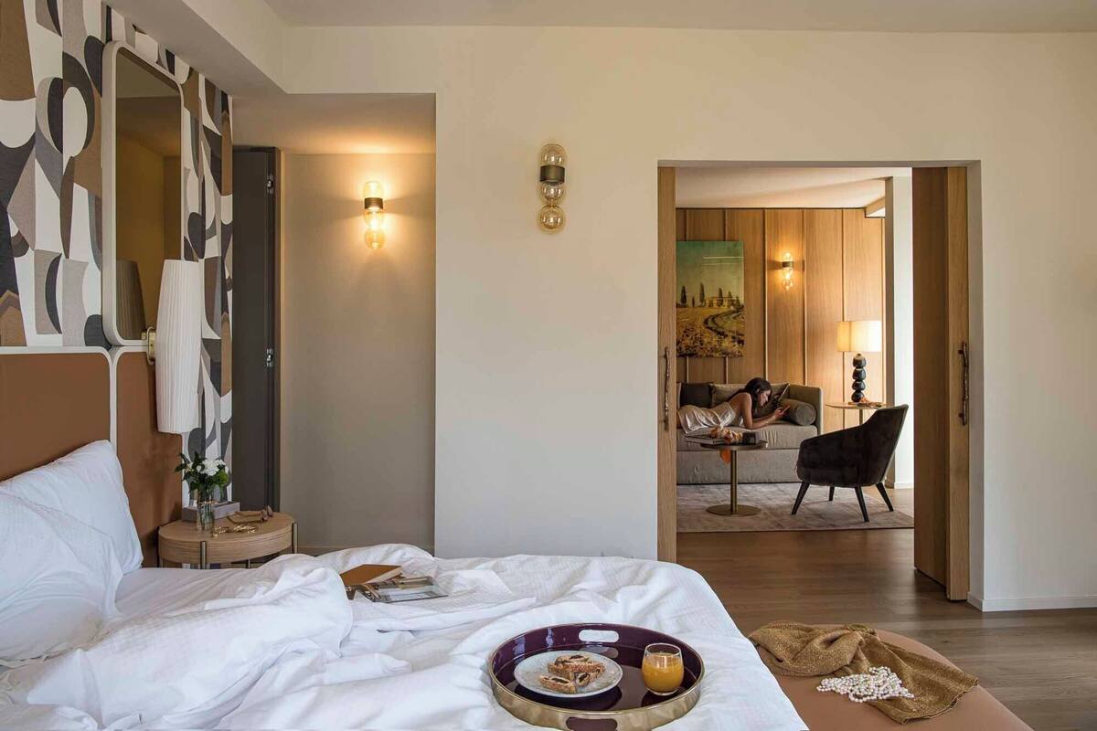 A hotel room with a bed, bedside table, lamp, and a view into the dining area.
