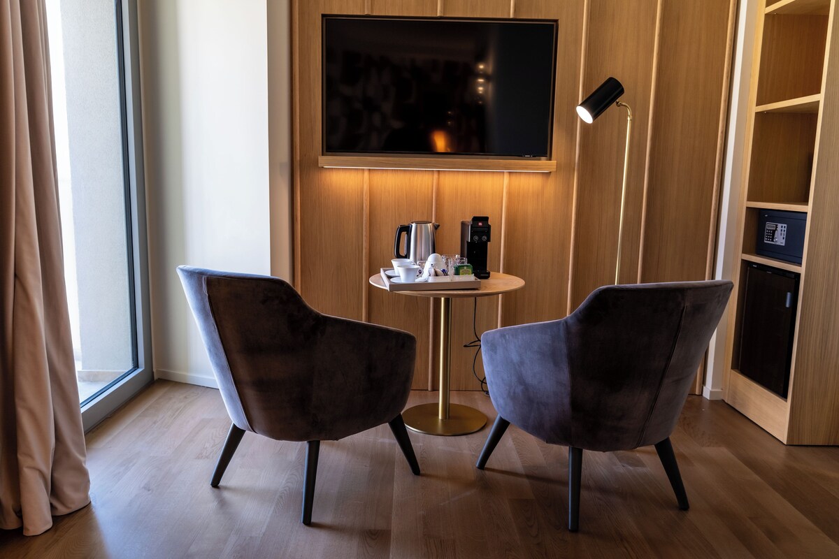 A modern room with two dark chairs, a small round table, a flat-screen TV mounted on the wall, and a floor lamp.