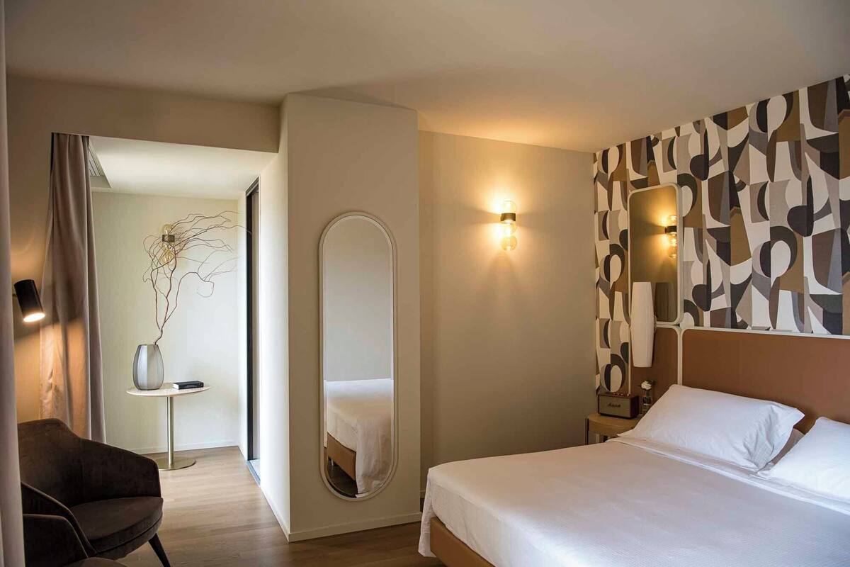 A hotel room with a bed, a round mirror, a wall-mounted light fixture, and a small table with a vase.